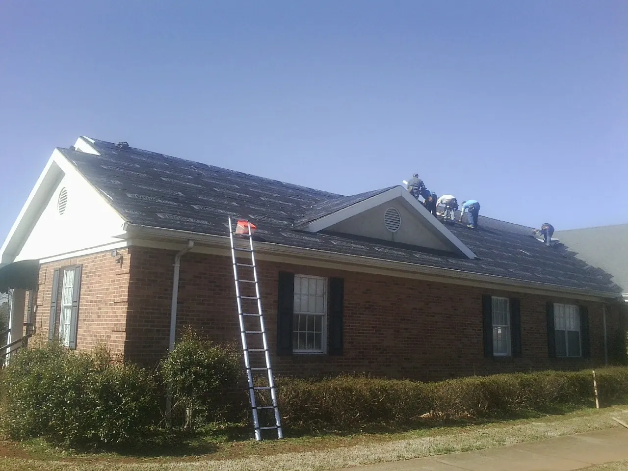 Crew tearing shingles off a brick ranch roof with a ladder set — Emergency Roof Repair work by our crew in Rotterdam Junction