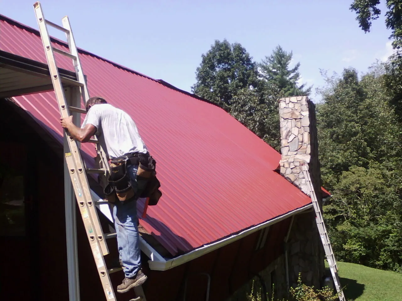 Roofer on a ladder beside a red metal roof and stone chimney — Soffit & Fascia Repair by Roofing Brothers in Rotterdam Junction